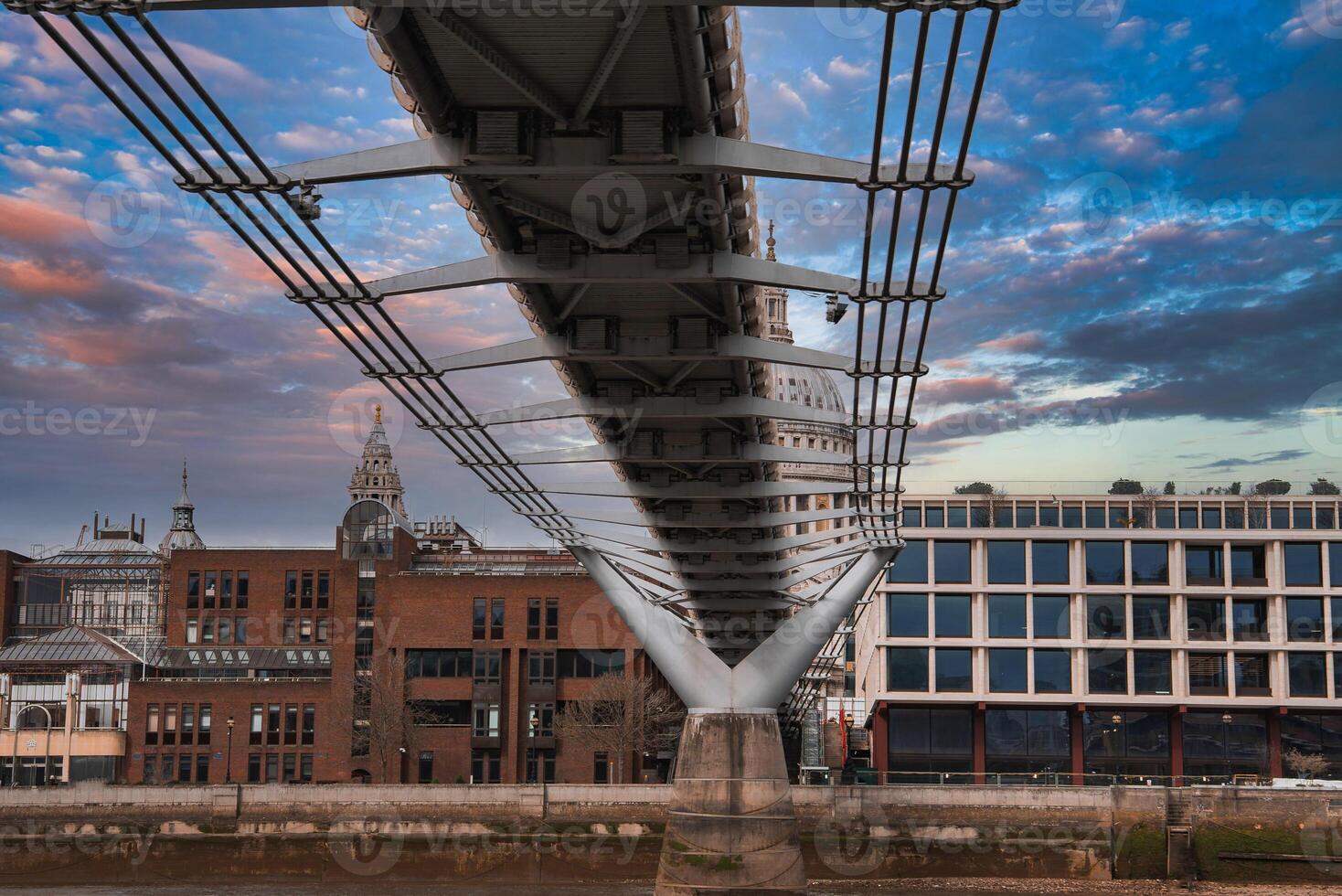 View of the Millennium Bridge's suspension cables and modern design, with St. Paul's Cathedral dome in the background under a colorful sky. photo