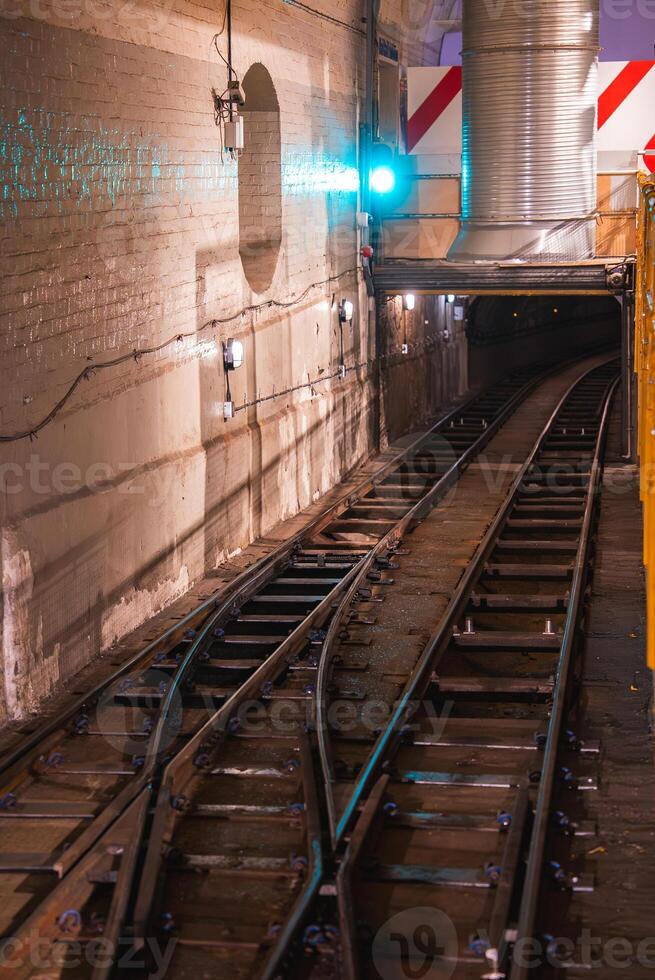Aged brick walls and dual railway tracks curve into the distance in a London metro tunnel. A green signal light glows, with ventilation ducts overhead. photo