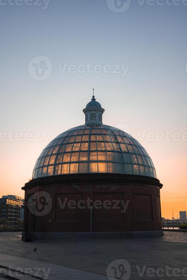 The domed glass and brick entrance of the Greenwich Foot Tunnel is lit by sunset hues. Modern buildings and the River Thames are visible in the background. photo