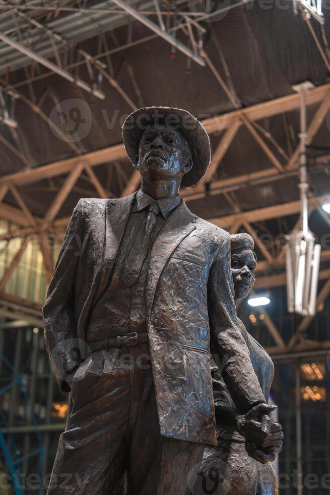 A bronze statue of a man in a suit and hat holding hands with a woman, set indoors against a steel and glass structure in Waterloo train station in London. photo