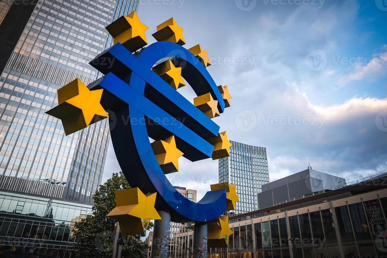 The Euro sculpture in Frankfurt, Germany, stands before modern skyscrapers, including the European Central Bank, with greenery and people nearby photo