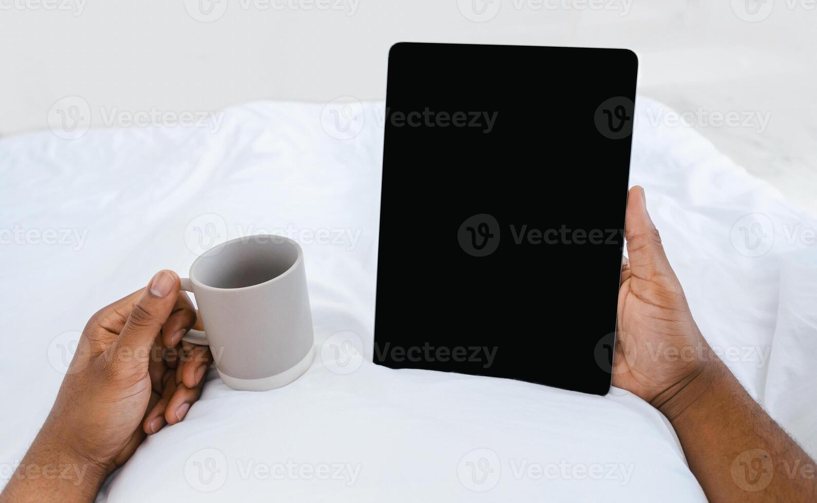 Black Male Hands Holding Digital Tablet With Empty Screen Reading E-Book Online Lying In Bed At Home, Enjoying Morning Coffee. Application Advertising, Weekend Leisure. Cropped, Mockup, POV photo