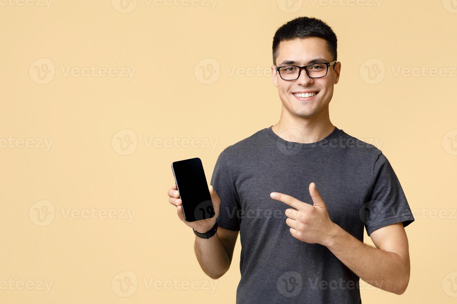 Blogger insists at better offer for customers. Happy handsome young guy in brown t-shirt and glasses, points finger at mobile phone with blank screen advertising app, isolated on beige background photo