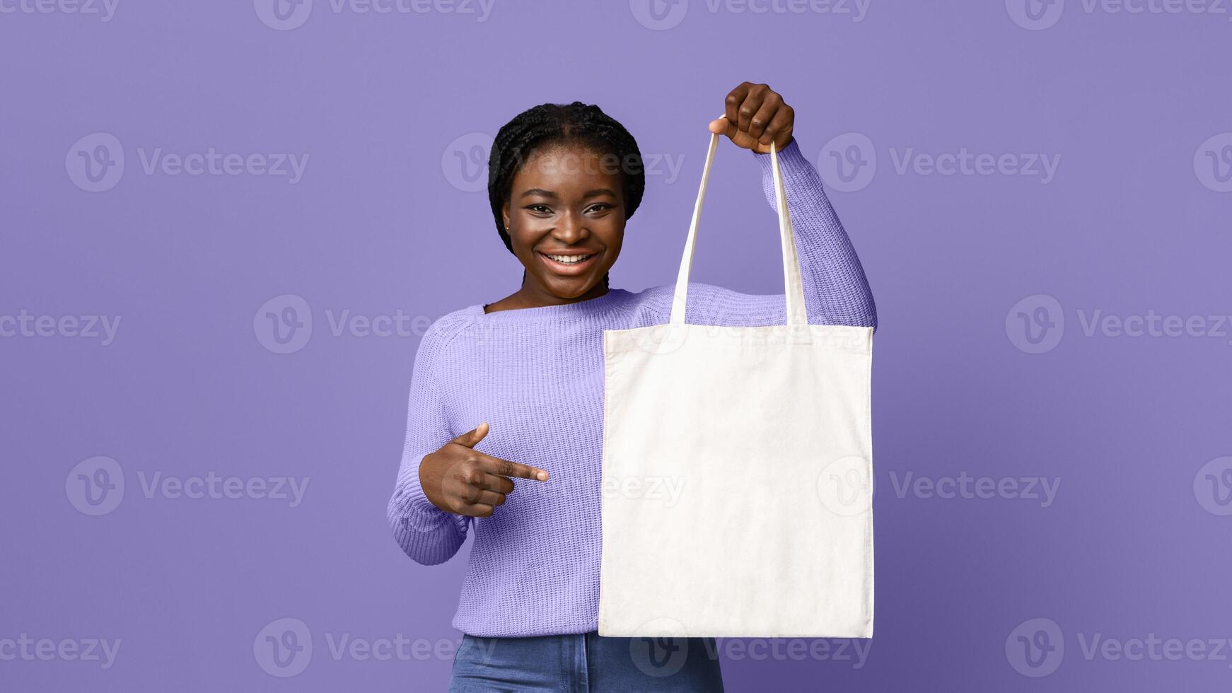 Cheerful Black Woman Holding And Pointing At Blank Textile Tote Eco Canvas Shopping Bag While Posing Over Purple Studio Background, Looking At Camera, Showing With Finger At Copy Space For Your Logo photo