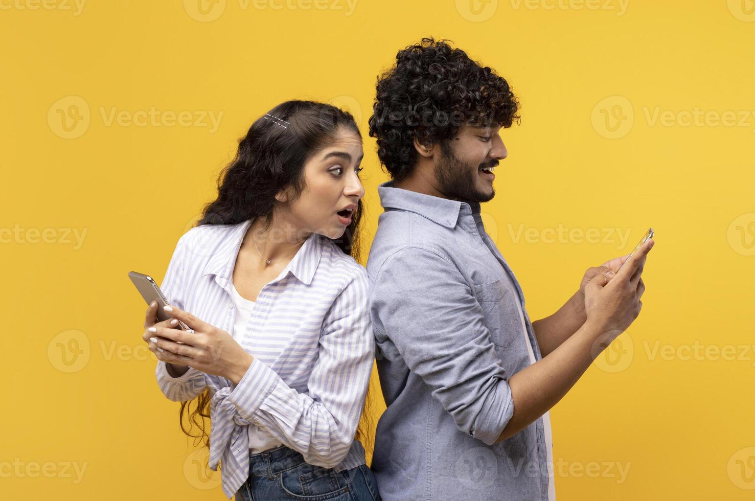 Shocked indian woman spying on her smiling boyfriend who using smartphone, chatting or scrolling social media news feed. Young couple standing back to back over yellow studio background photo