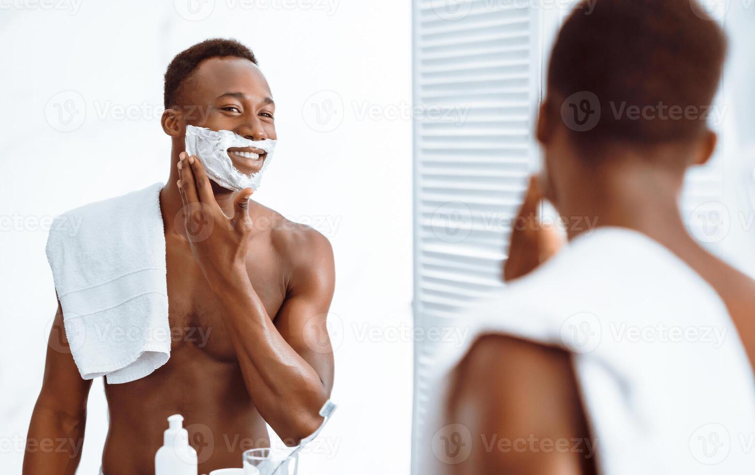 Shirtless African Guy Applying Shave Foam On Face Preparing For Shaving Standing Near Mirror In Bathroom Indoors. Male Facial Skincare And Selfcare Routine Concept. Selective Focus photo