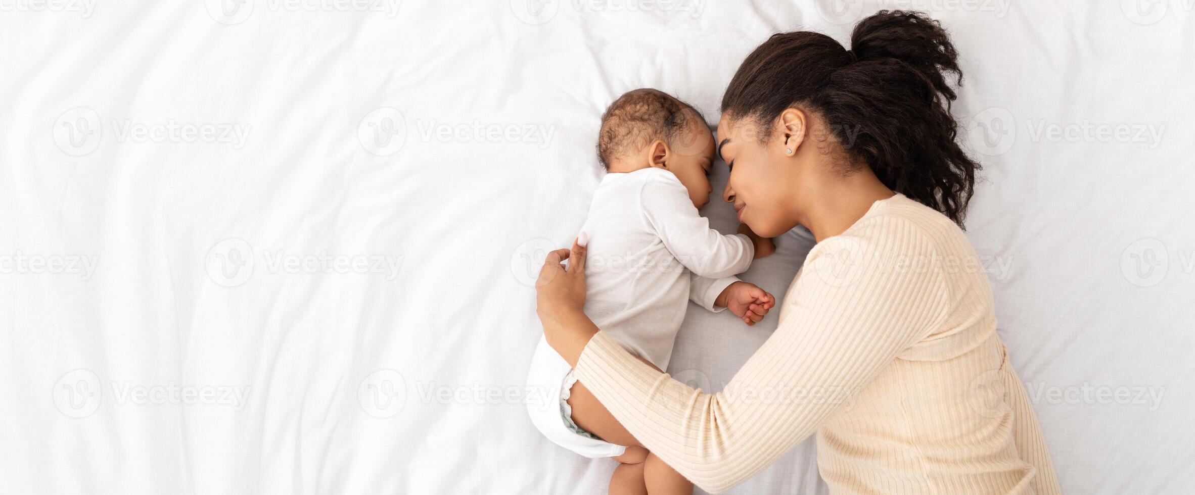 African Mother Hugging Sleeping Baby Lying In Bed Indoor, High Angle Shot. Loving Mom And Toddler Infant Napping Resting During Daytime Sleep. Panorama, Free Space For Text photo