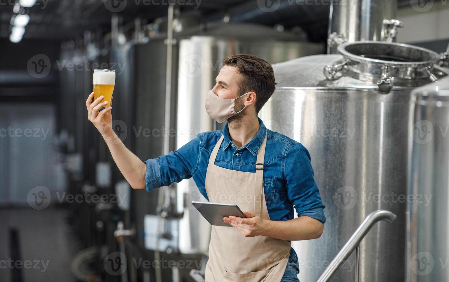Beer quality control. Man in apron and protective mask with digital tablet looking at glass of beer on brewery factory, free space photo