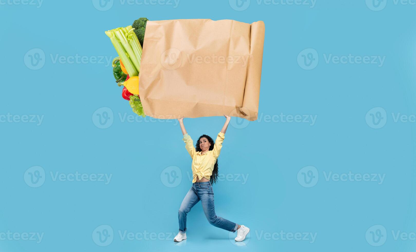 A shopper proudly holds a large paper bag filled with vibrant vegetables and fruits. The backdrop features a bright blue color, emphasizing the excitement of grocery shopping. photo