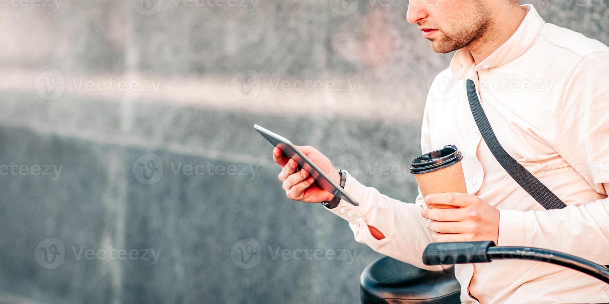 Work and study online, urgent task and eco transport. Serious handsome millennial man sits on bicycle with cup of takeaway coffee and reads message on tablet, on gray street wall background, outdoor photo