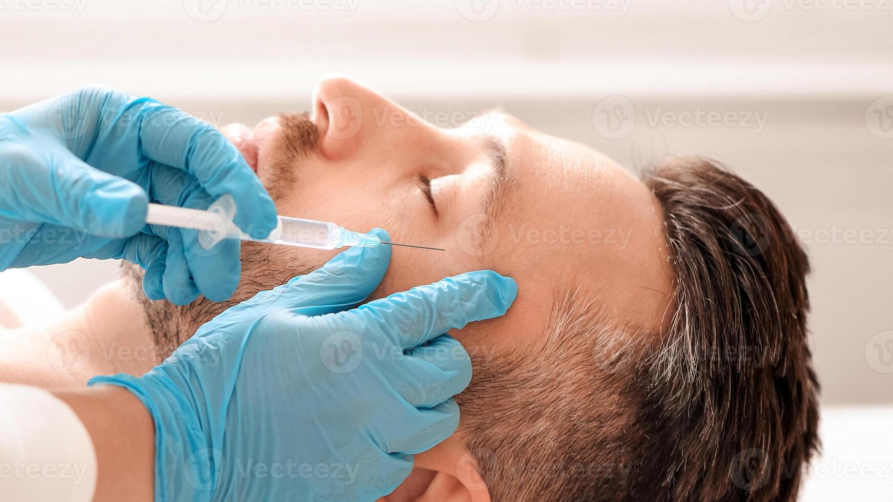 Cosmetologist making eye lifting procedures for handsome middle aged man at aesthetic clinic, side view. Man getting beauty injection against eye wrinkles from unrecognizable plastic surgeon photo