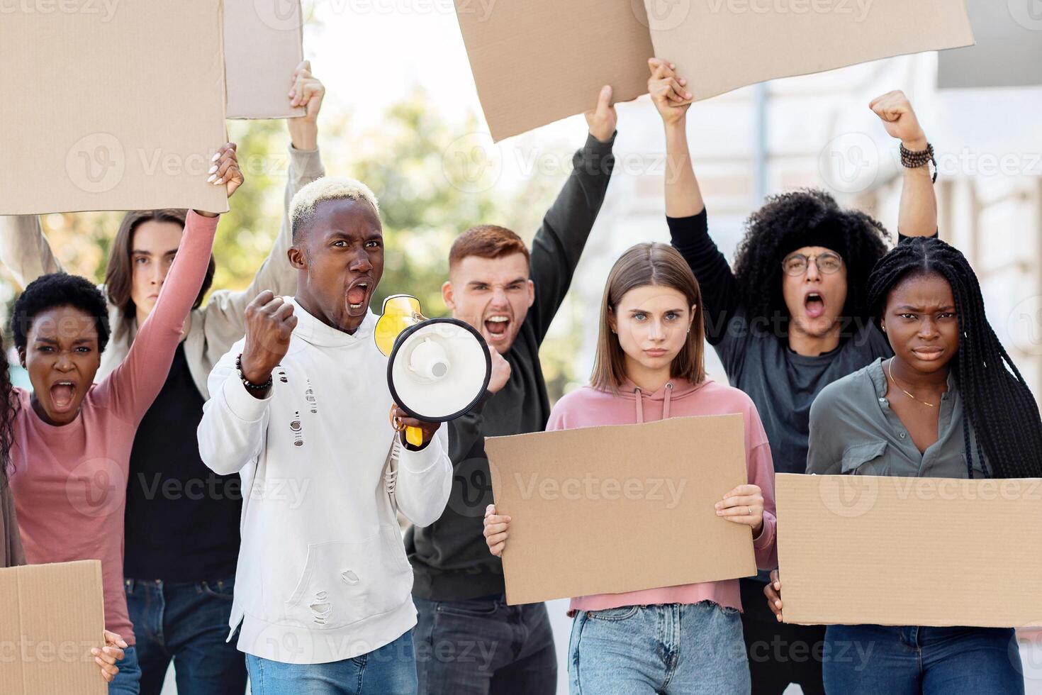 Furious multiracial students with empty placards having strike or demonstration on the street. Motivated african american man with loudspeaker chanting slogans over demonstrating crowd outdoors photo