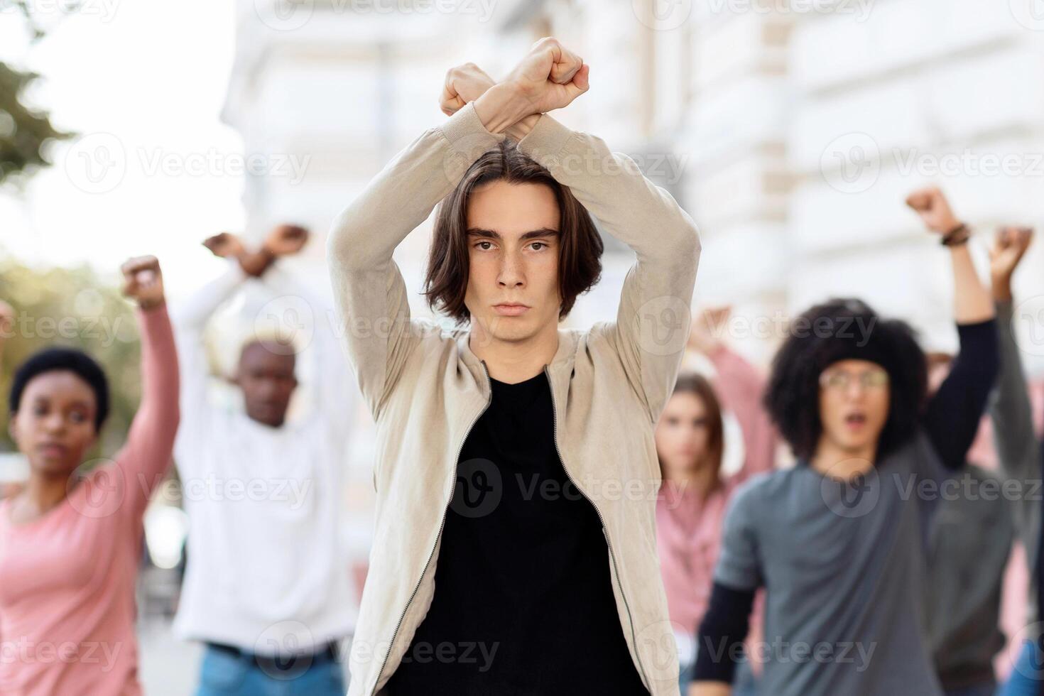 Young man leading multiracial group of activists, crossing fists up, showing unity and strength, millennial strikers different nationalities walking by street, making riot against quarantine rules photo