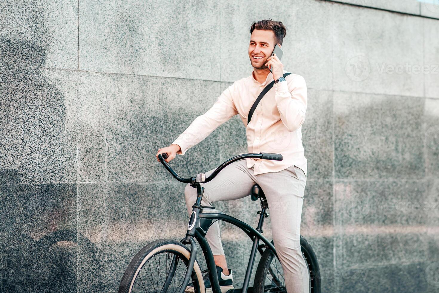 Urgent call, work on move and active, sports lifestyle. Smiling attractive millennial guy manager speaks on phone and rides on bike at city, on gray street wall background, outdoor, free space photo