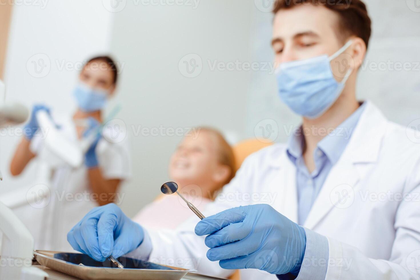 Prepare of instruments for procedure of dental treatment and examination. Focus on serious doctor millennial in coat, protective mask and gloves with girl patient and nurse on blurred background photo