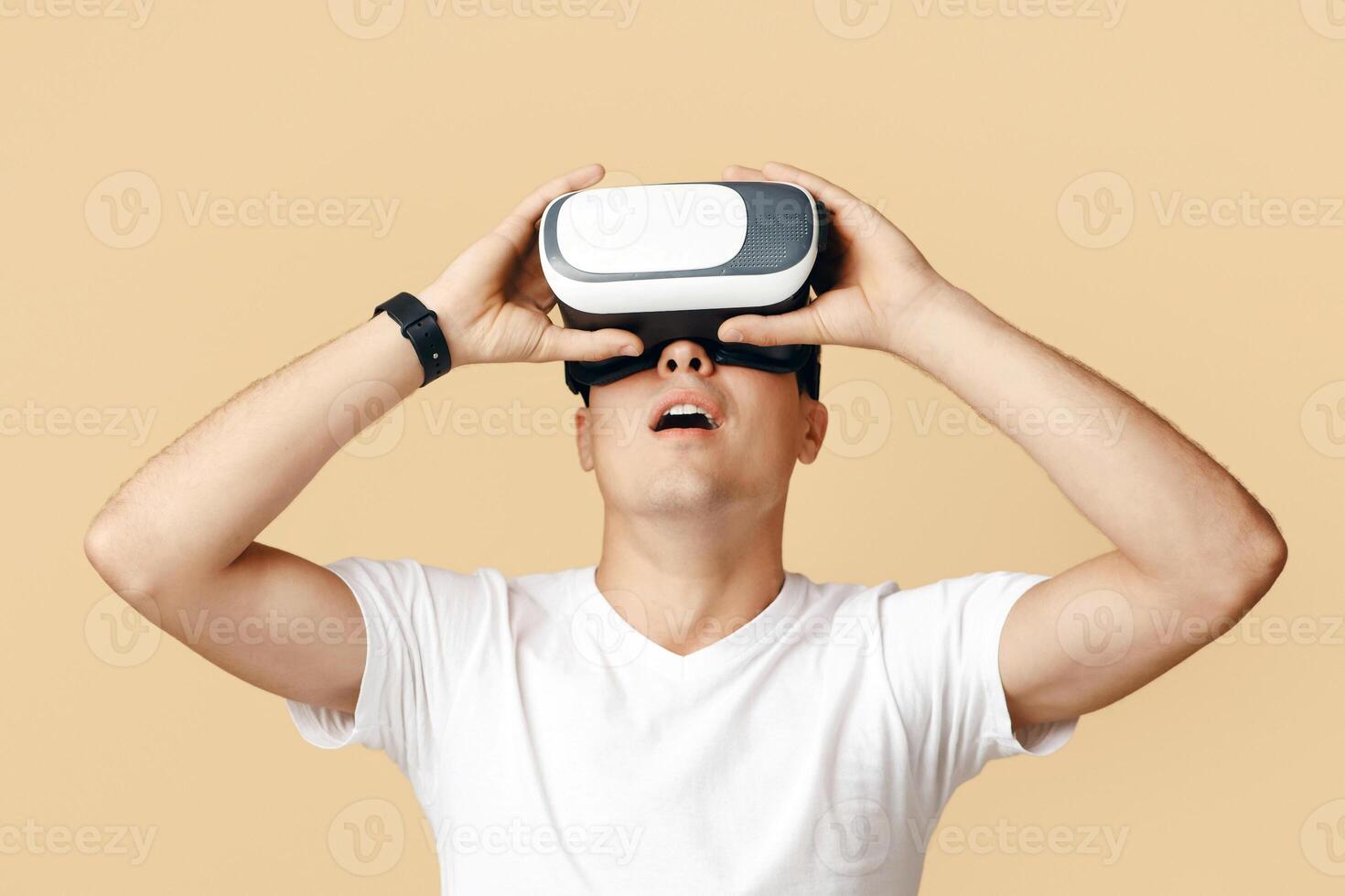 Amazing virtual world and modern technology for study and entertainment. Surprised young man with open mouth in virtual reality glasses looking up at empty space, isolated on beige background photo