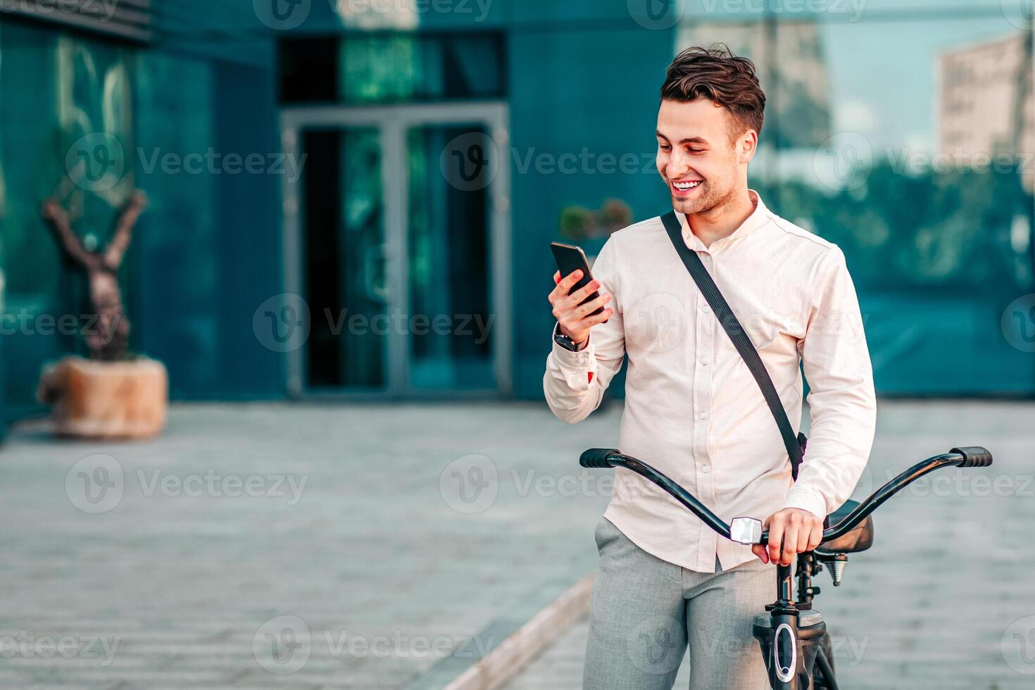 Outdoor portrait of handsome young man with mobile phone and bicycle in street. Man hipster manager in stylish clothes reads good message from client about deal near city office, outdoor, free space photo