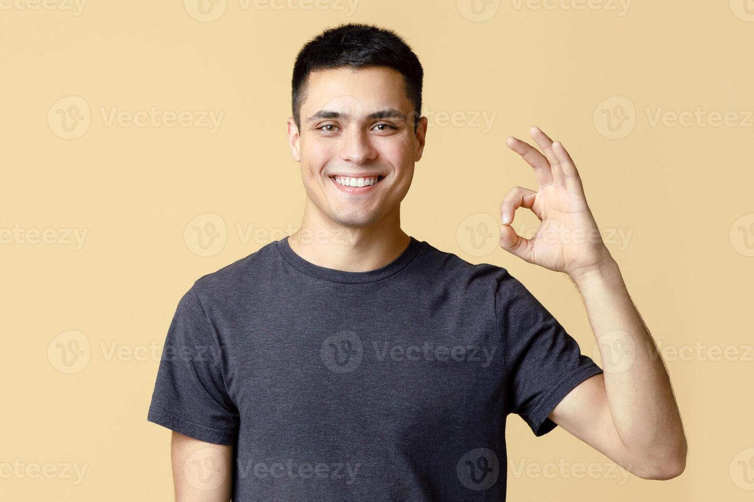Great offer, result and review from blogger. Handsome young cheerful student man in brown t-shirt shows okay gesture symbol with hand, isolated on beige background, studio shot, free space photo