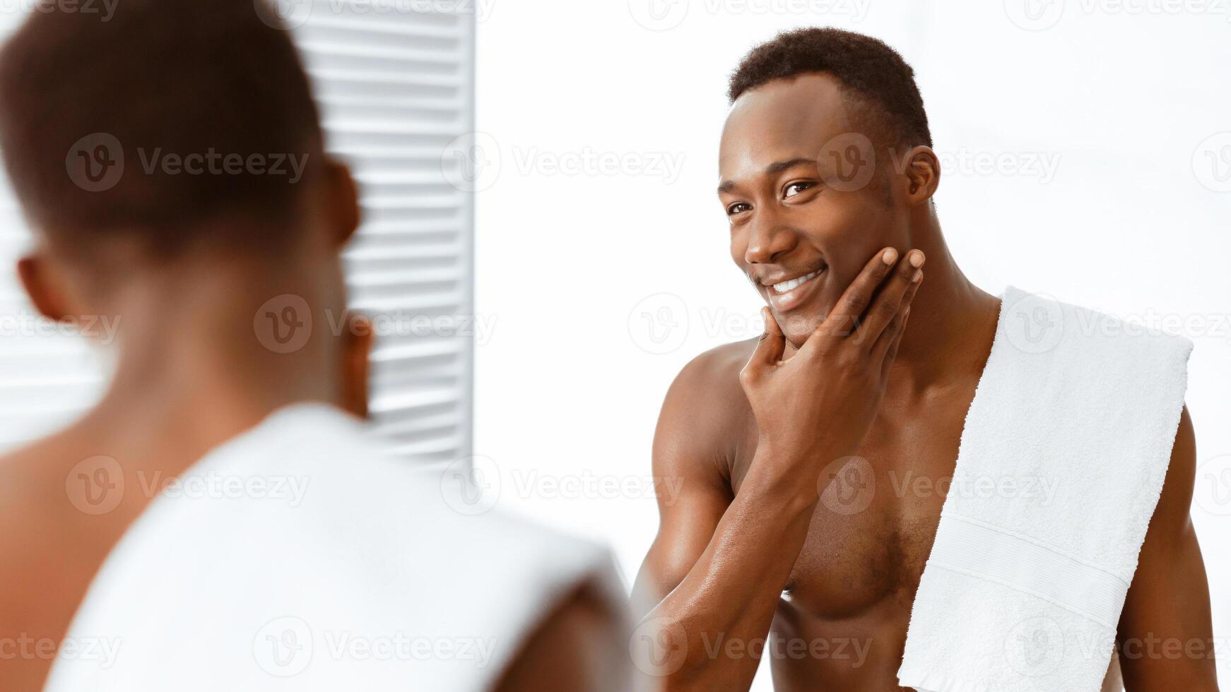 Happy Well-Shaved Black Guy Touching Smooth Face Caring For Skin After Shaving Routine Standing Near Mirror In Modern Bathroom At Home. Male Facial Skincare And Self-Care Concept. Panorama photo