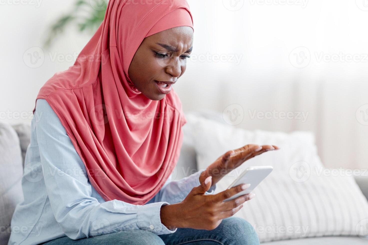 Bad Message. Annoyed Black Muslim Woman In Hijab Shouting At Cellphone In Her Hands, Sitting On Couch At Home, Received Unpleasant Sms, Complaining About Something, Having Phone Quarrel, Free Space photo