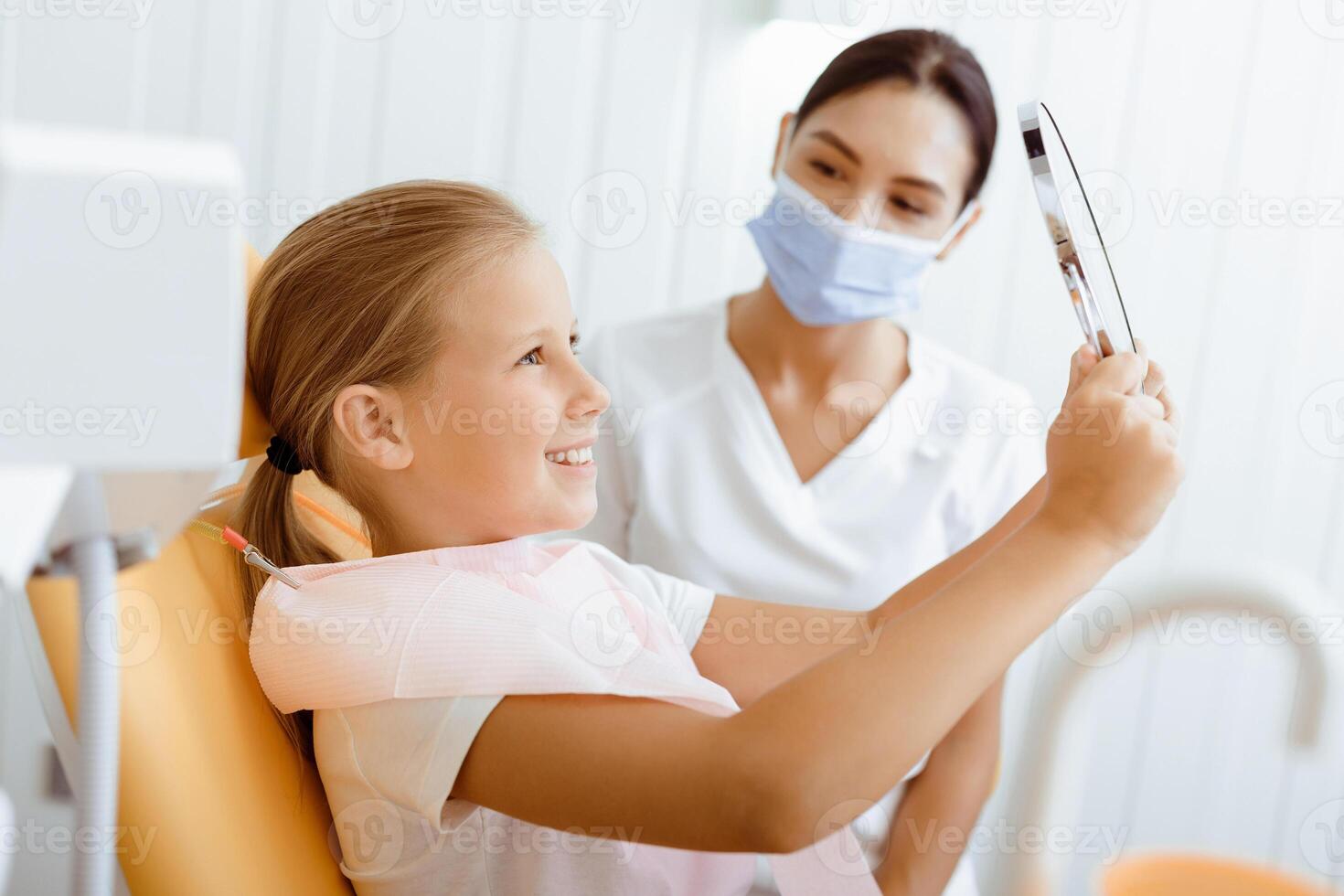 Great dentist work and perfect kids teeth. Happy little girl in medical chair looks in mirror, young female doctor in protective mask and uniform looks at baby in modern clinic at daylight, free space photo