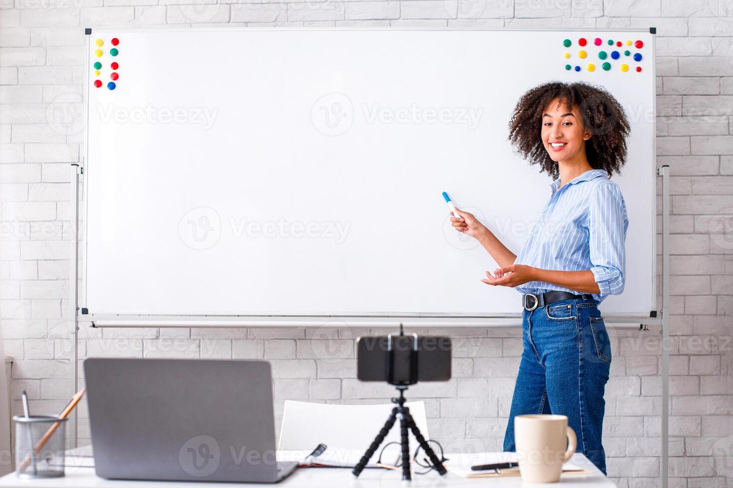 Modern english lessons at home. Smiling african american teacher points to white board with rules and records for students in living room interior with laptop and smartphone, free space photo