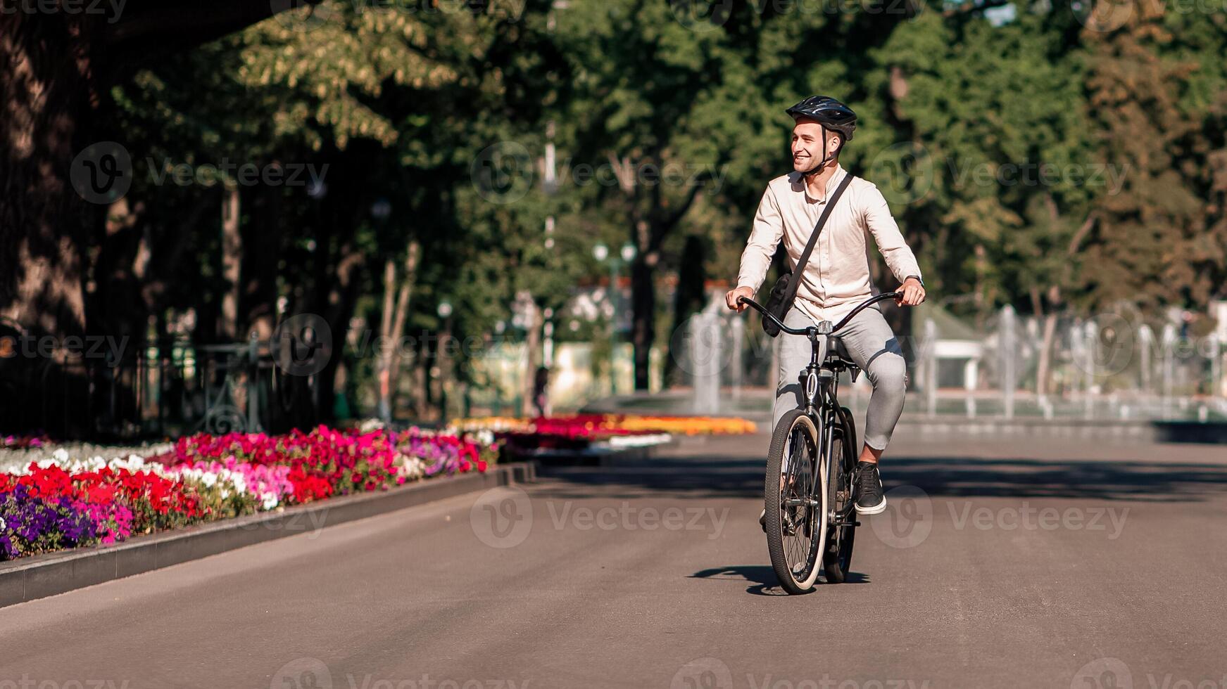Businessman enjoying the trip to work in morning. Happy millennial guy employee in protective helmet rides on bicycle in city in summer park, and look at landscape with fountain, outdoor, free space photo