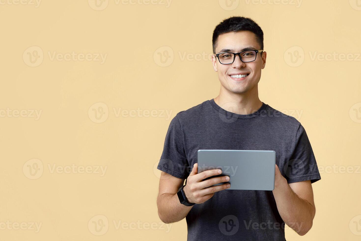 Modern work, social media and online communication. Attractive happy young man student in glasses, with smart watch typing in digital tablet, isolated on beige background, studio shot, free space photo
