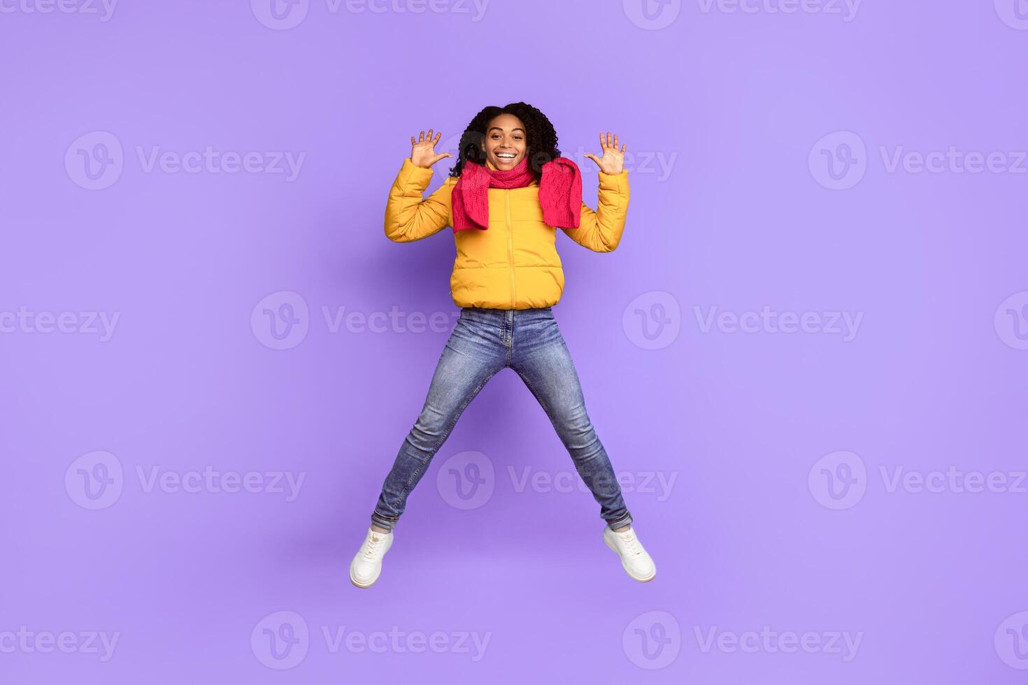 Hello. Joyful African American Woman Jumping Waving Hands In Mid-Air And Smiling To Camera Posing Wearing Warm Jacket Over Blue Studio Background. Winter Advertisement Banner. Full Length Shot photo