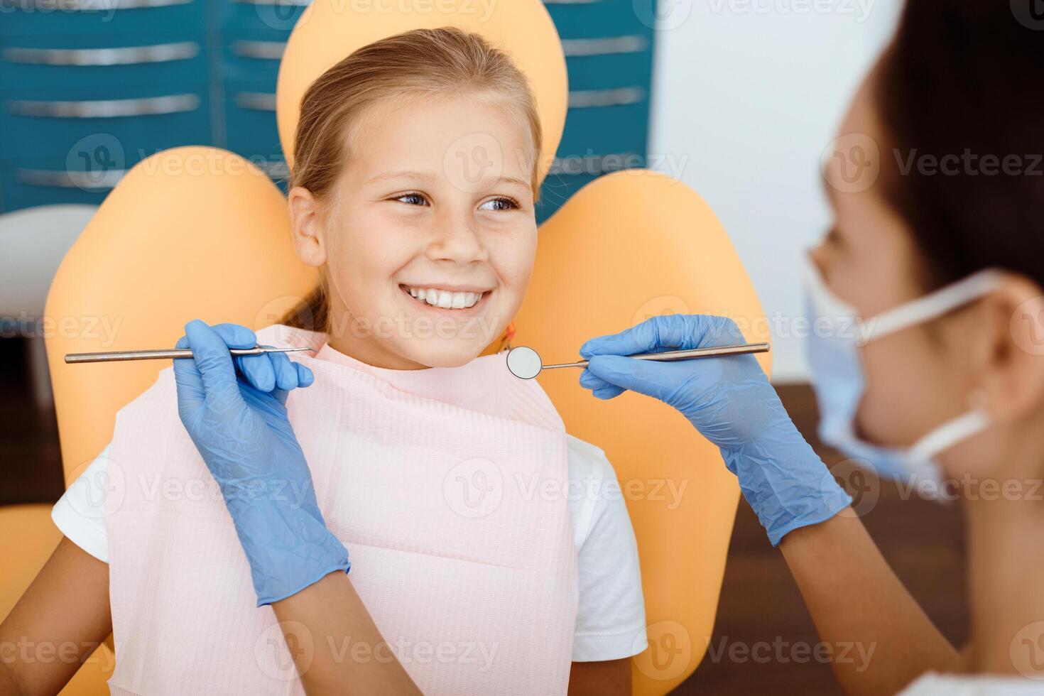 Routine check-ups, snow-white smile and pediatric dentistry. Young female in protective mask and gloves examination happy little kid in medical chair, looking at doctor with tools in hand, cropped photo