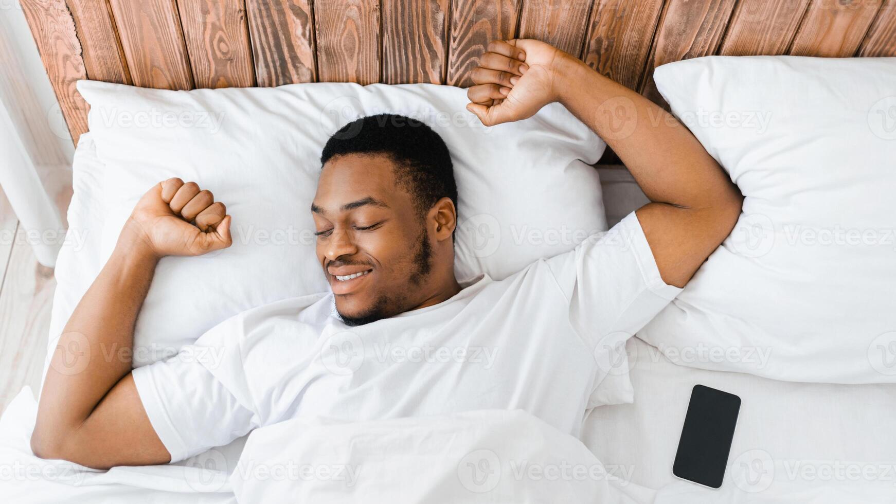 Good Morning Concept. Wellslept Black Guy Awakening In Bed Stretching Hands After Sleeping In Bedroom At Home. Man Waking Up In Bed, Recreation And Rest. Panorama, High-Angle View photo