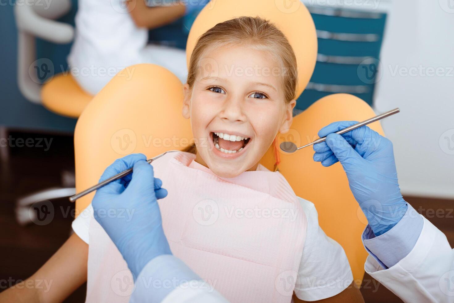 Great visit to dentist. Happy little child with napkin on chest sits in chair and looks at camera, doctor in rubber gloves with medical instruments ready for teeth procedure, close up, pov photo