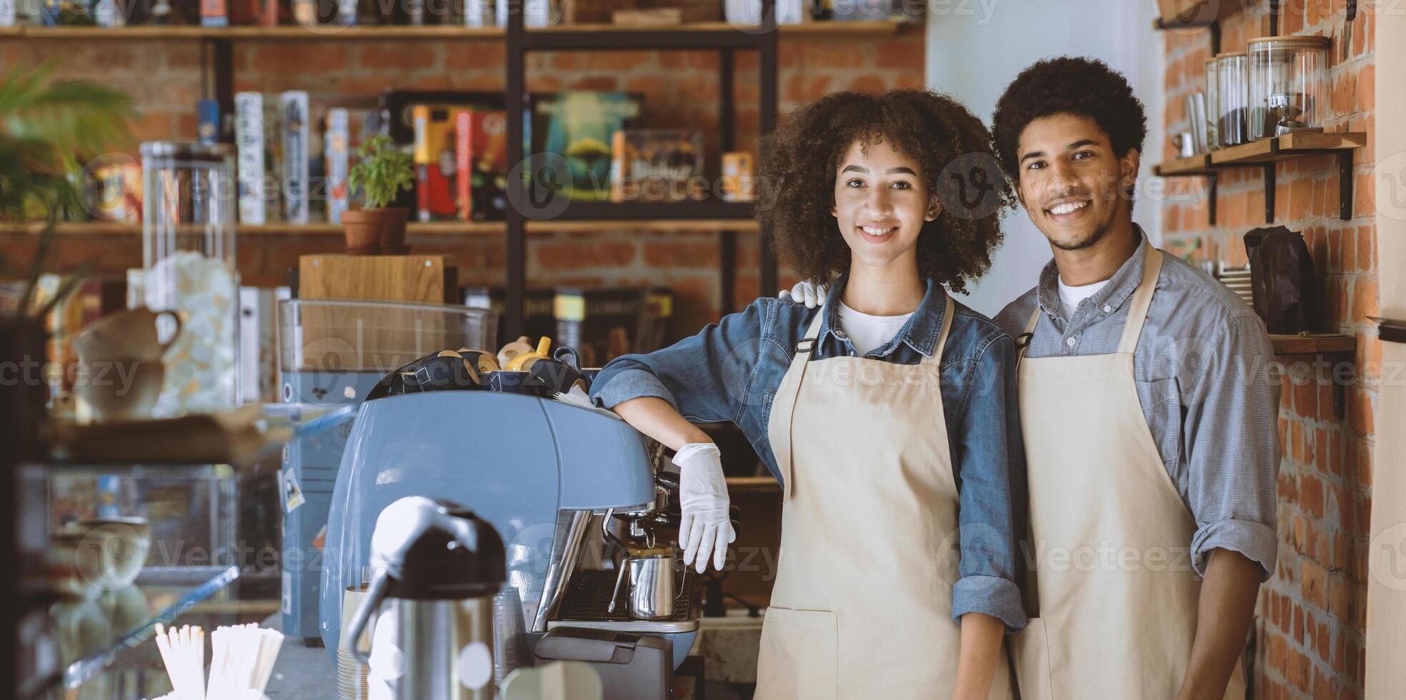 Meeting and waiting for clients in modern cafe after lockdown. Smiling millennial african american guy and lady in aprons and gloves with espresso machine and equipment in morning in cafeteria photo