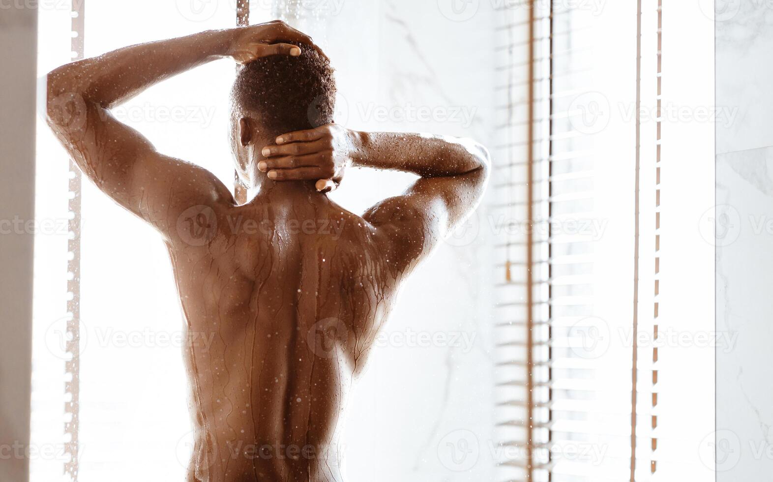 Unrecognizable Black Man Taking Shower Standing Back To Camera Washing Body Under Falling Water In Modern Bathroom At Home. Male Bodycare Concept. Rear View, Blank Space For Text photo