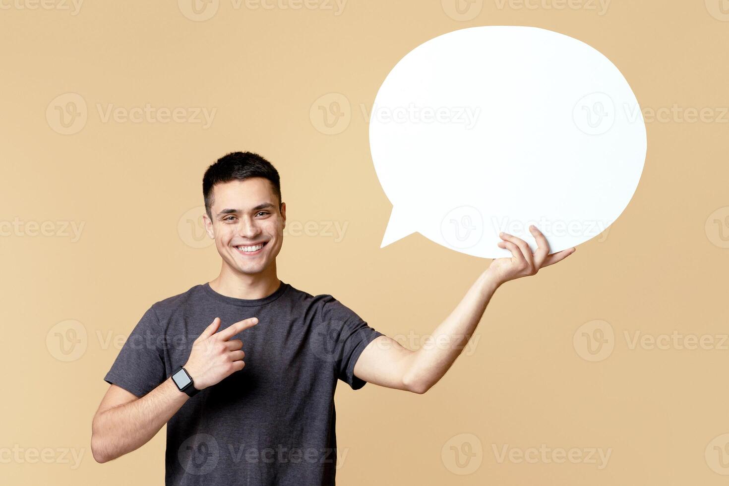 Cool cheerful guy in grey t-shirt holding empty blank white speech bubble and pointing at it, isolated on beige studio background, copy space photo