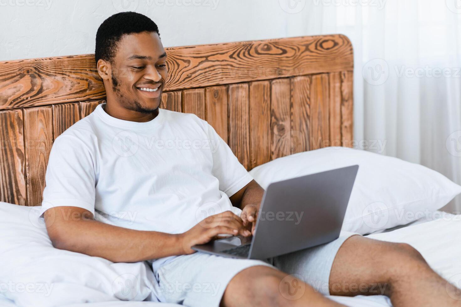 Happy Black Freelancer Guy Working On Laptop Distantly Online Browsing Internet Sitting In Bed In Bedroom At Home. Remote Computer Job And Freelance Career Concept photo