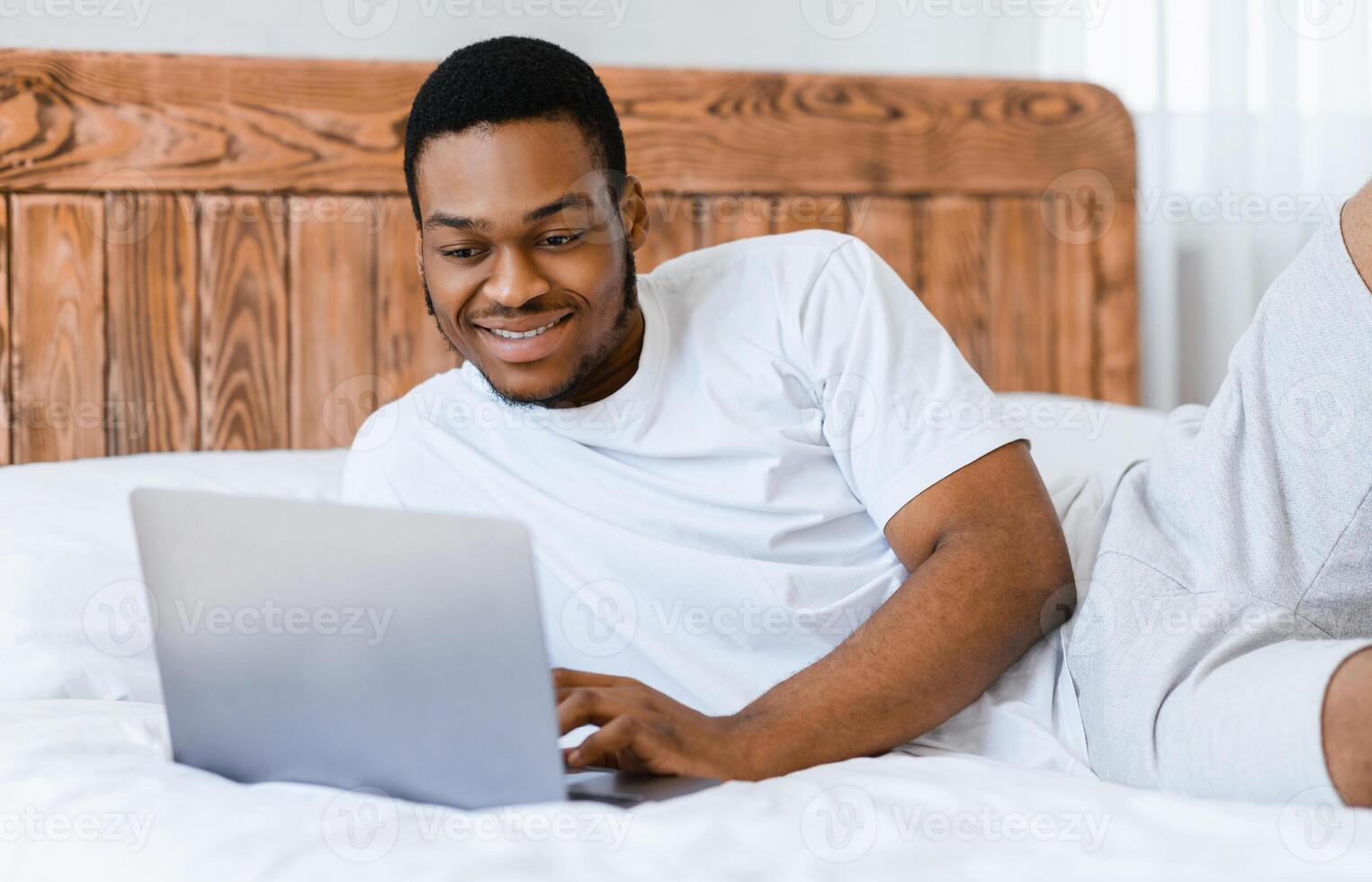 Happy African American Freelancer Man Working On Laptop Computer Browsing Internet Lying In Bed At Home. Remote Job, Freelance Career And Lifestyle Concept. Selective Focus photo