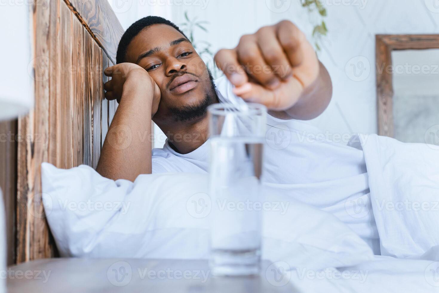Sick African Guy Taking Medicine, Putting Pill Into Glass Of Water Lying In Bed In Bedroom At Home. Flu And Cold Medication Treatment Concept. Selective Focus photo