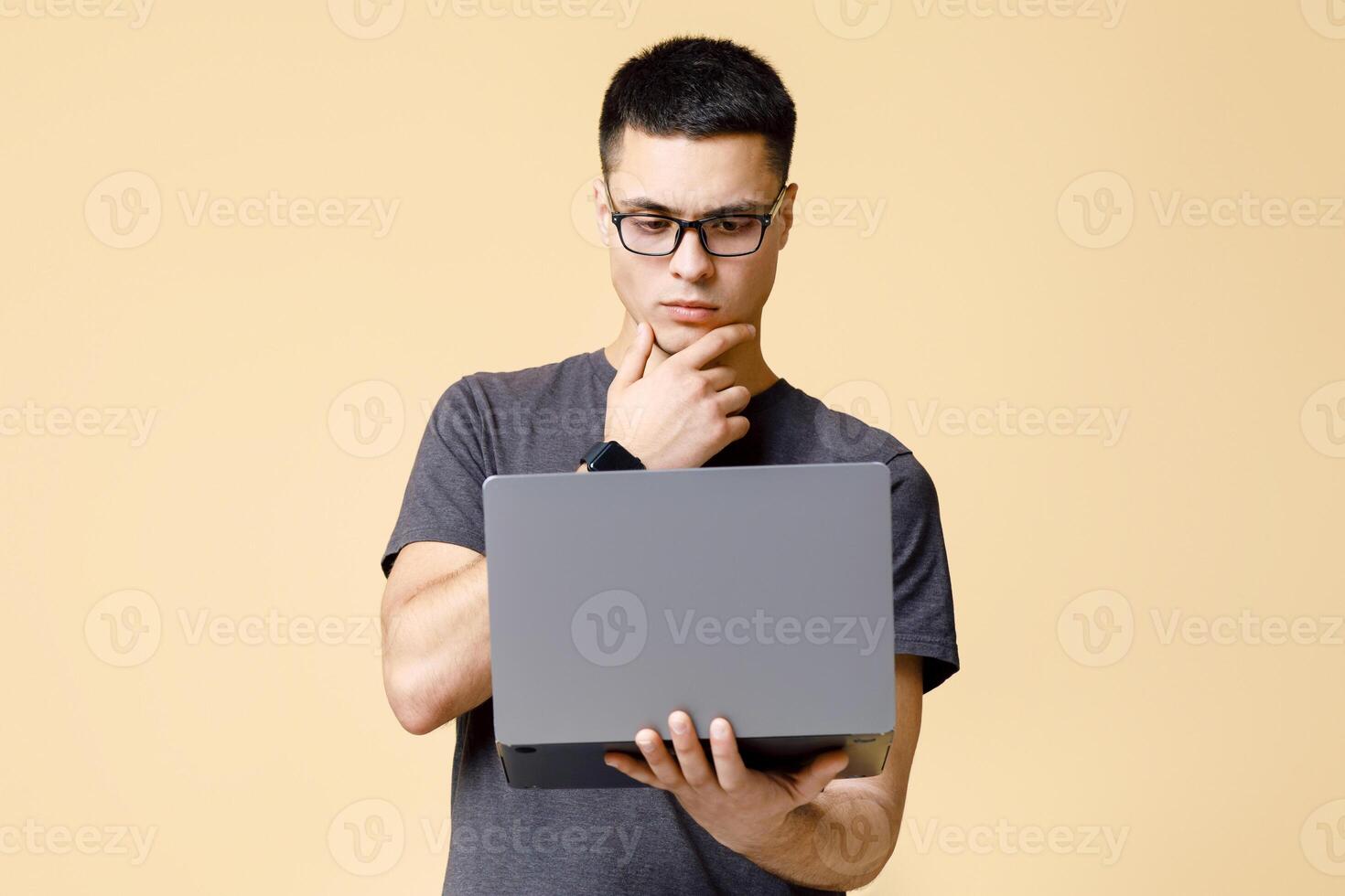 Designer and freelancer works online. Pensive serious young man in brown t-shirt rubs his chin and looks at laptop, learning information or task, isolated on beige background, studio shot, free space photo