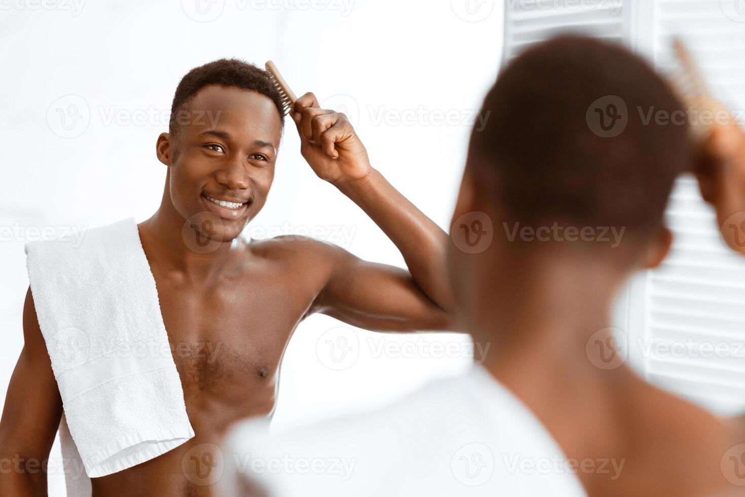 Shirtless African Man Brushing Hair Doing Haircare Routine Using Hairbrush Standing Near Mirror In Modern Bathroom At Home. Everyday Male Hairstyling Concept. Selective Focus photo