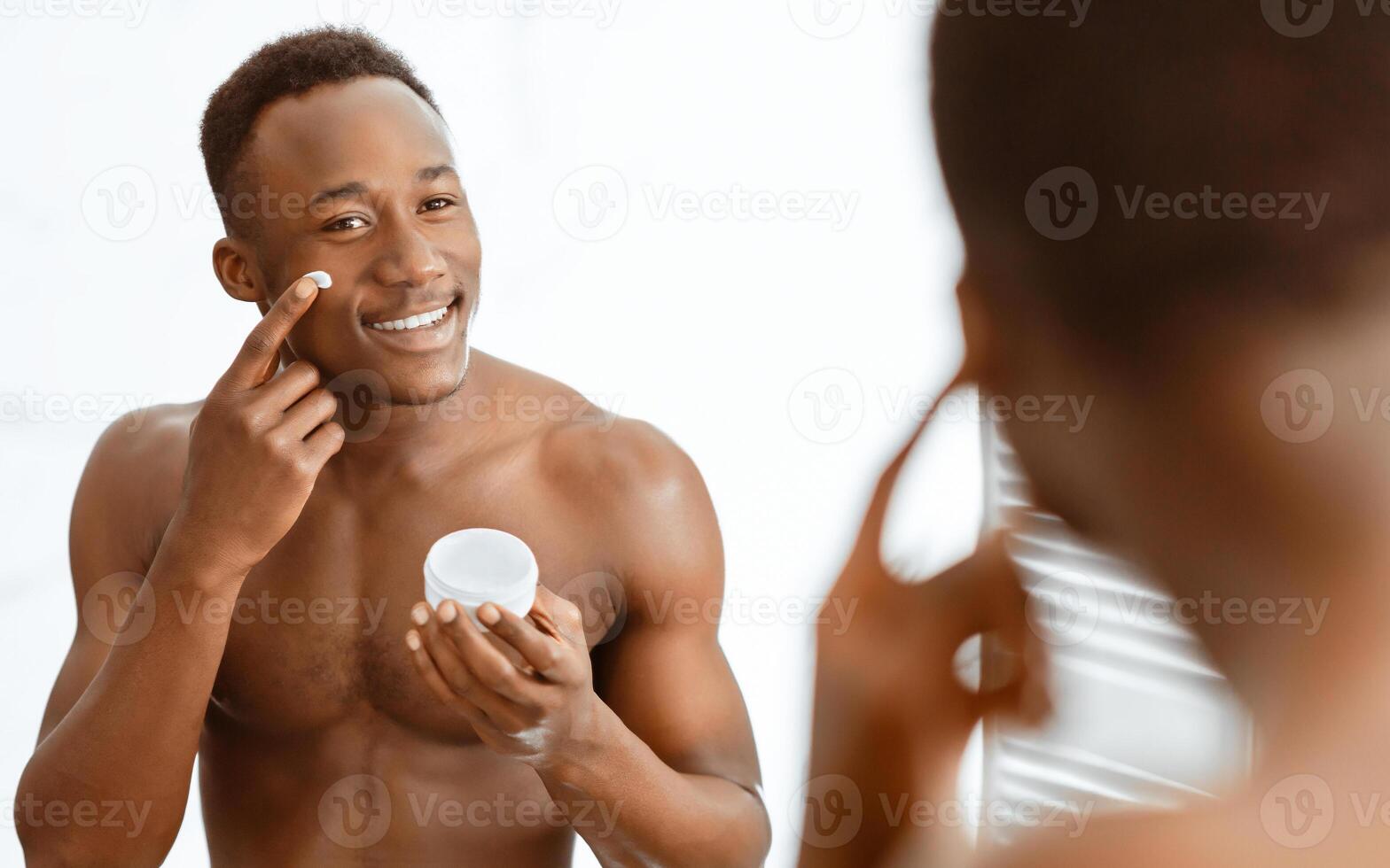 Facial Skincare. African American Man Applying Moisturizing Cream On Face Standing Looking At His Reflection In Bathroom Mirror At Home. Male Beauty And Skin Care Routine Concept. Selective Focus photo