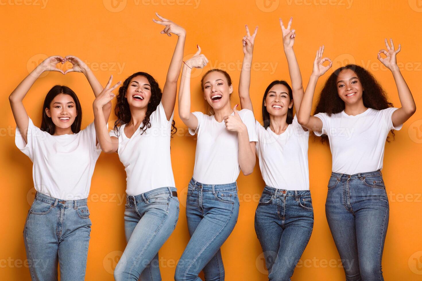 Group Of Multiracial Ladies Gesturing Different Positive Signs And Symbols Raising Hands Posing Over Yellow Studio Background, Smiling To Camera. Females Showing Okay, V-Sign And Thumbs Up Gestures photo