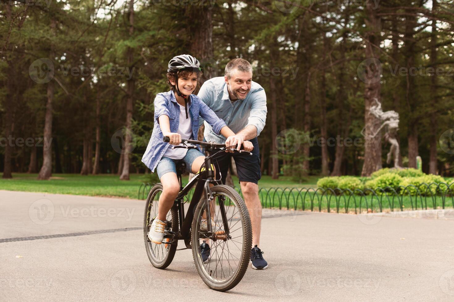 Leisure Concept. Portrait of cheerful adult father teaching his son how to ride a bicycle at public park, happy loving family spending time together on weekend, having fun outdoors in the summer photo