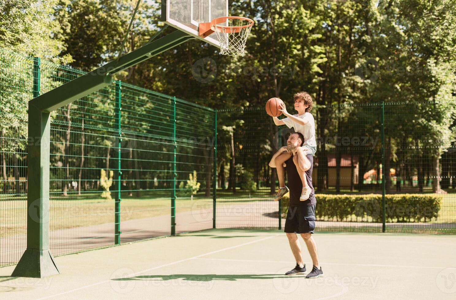 Assistance Concept. Man holding his son on shoulders, helping him to score a basket on playing field photo