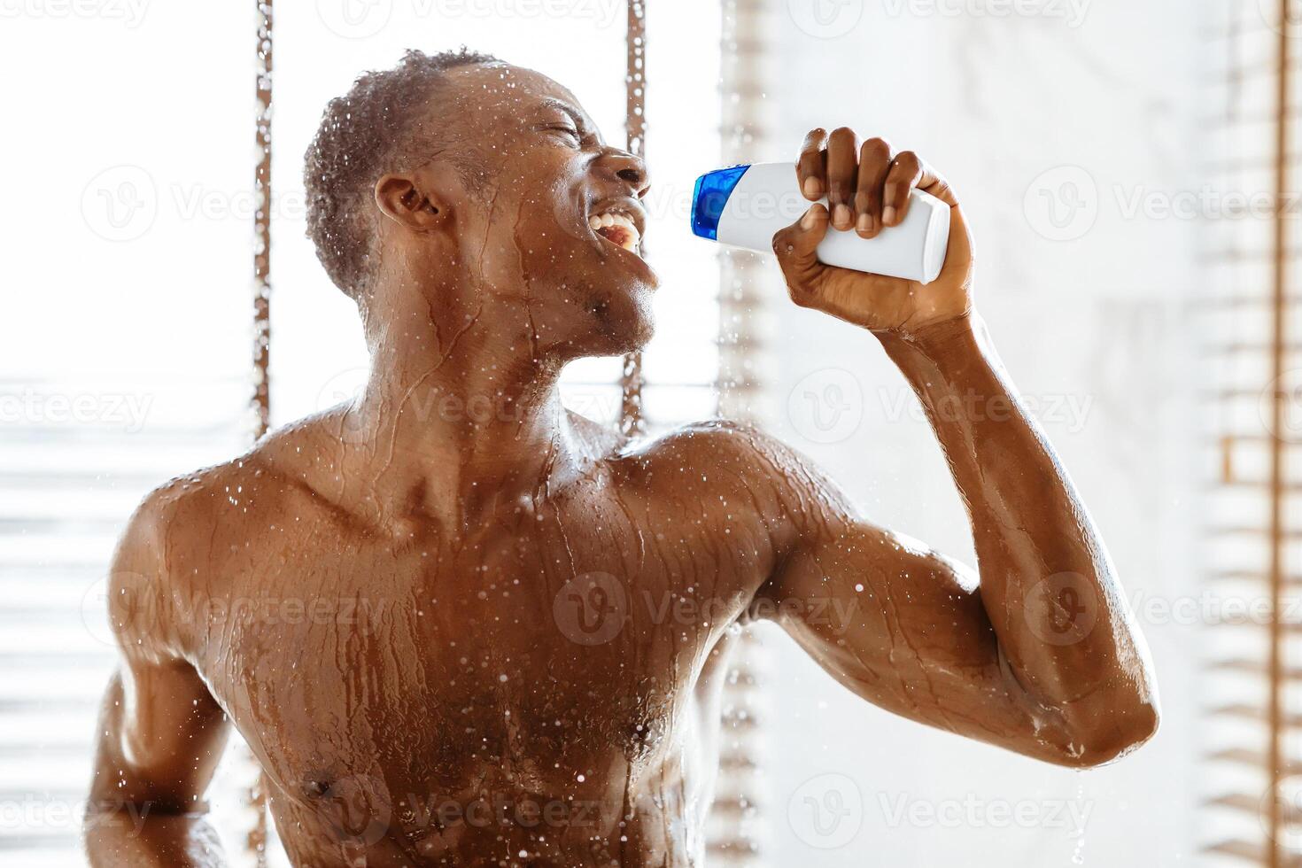 Positive african man singing taking shower holding shampoo bottle like microphone, standing under falling water drops in modern bathroom at home. Everyday hygiene, cleanliness and relaxation photo