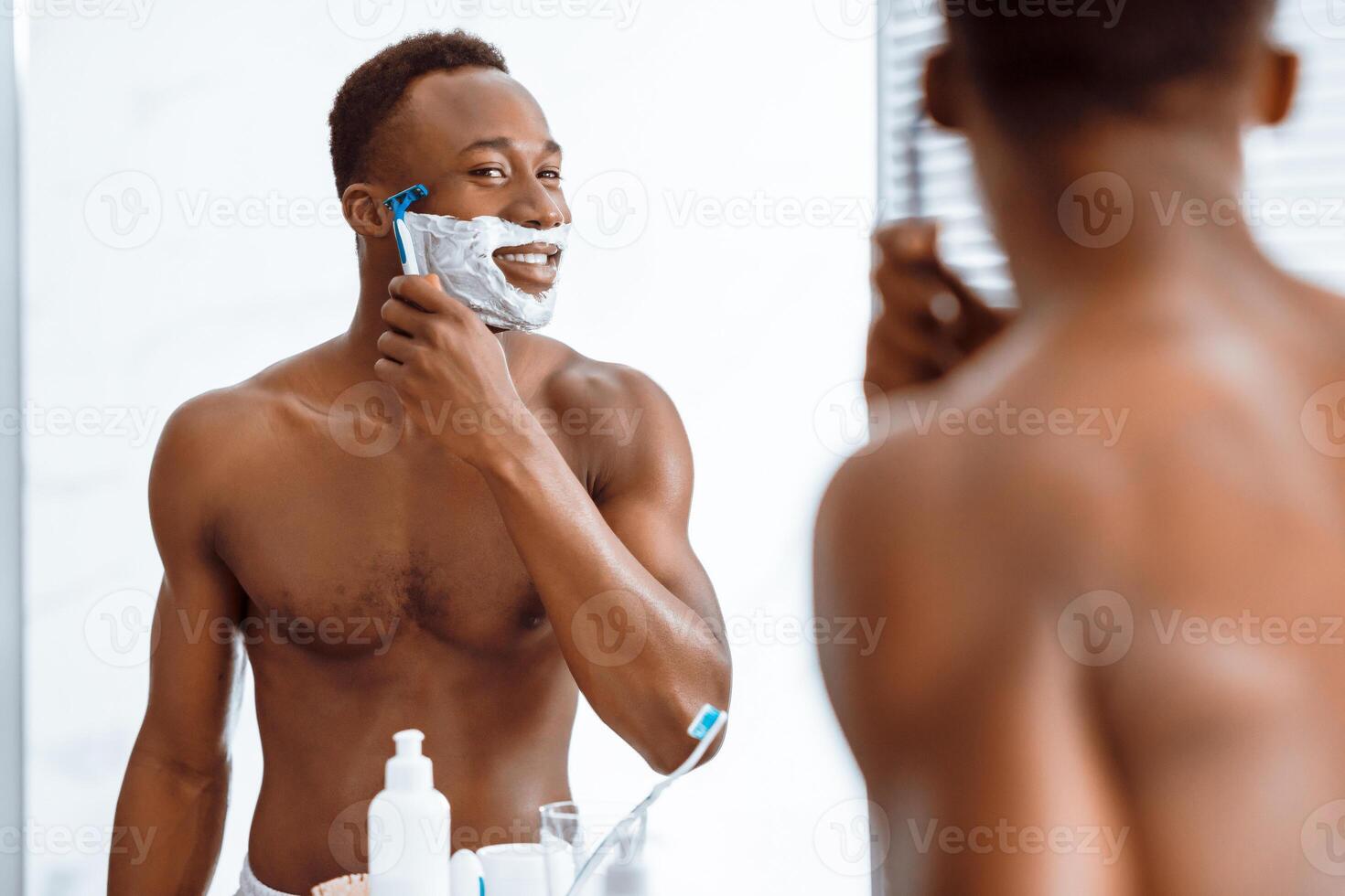Shirtless African Man Shaving Face Covered With Shave Foam Using Razor Standing Near Mirror In Bathroom At Home In The Morning. Male Facial Skincare Beauty Routine. Selective Focus photo