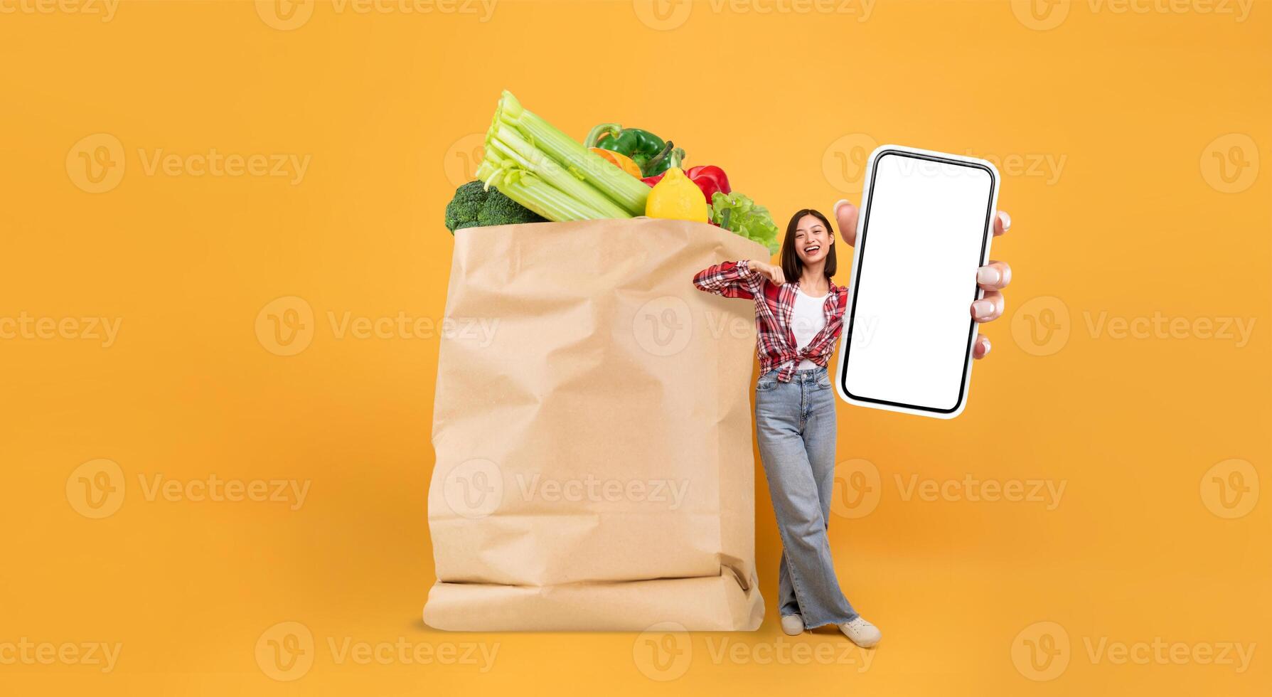 A shopper stands beside a large paper bag filled with fresh vegetables and fruits while holding a smartphone. This depicts modern grocery purchasing trends amidst changing habits. photo