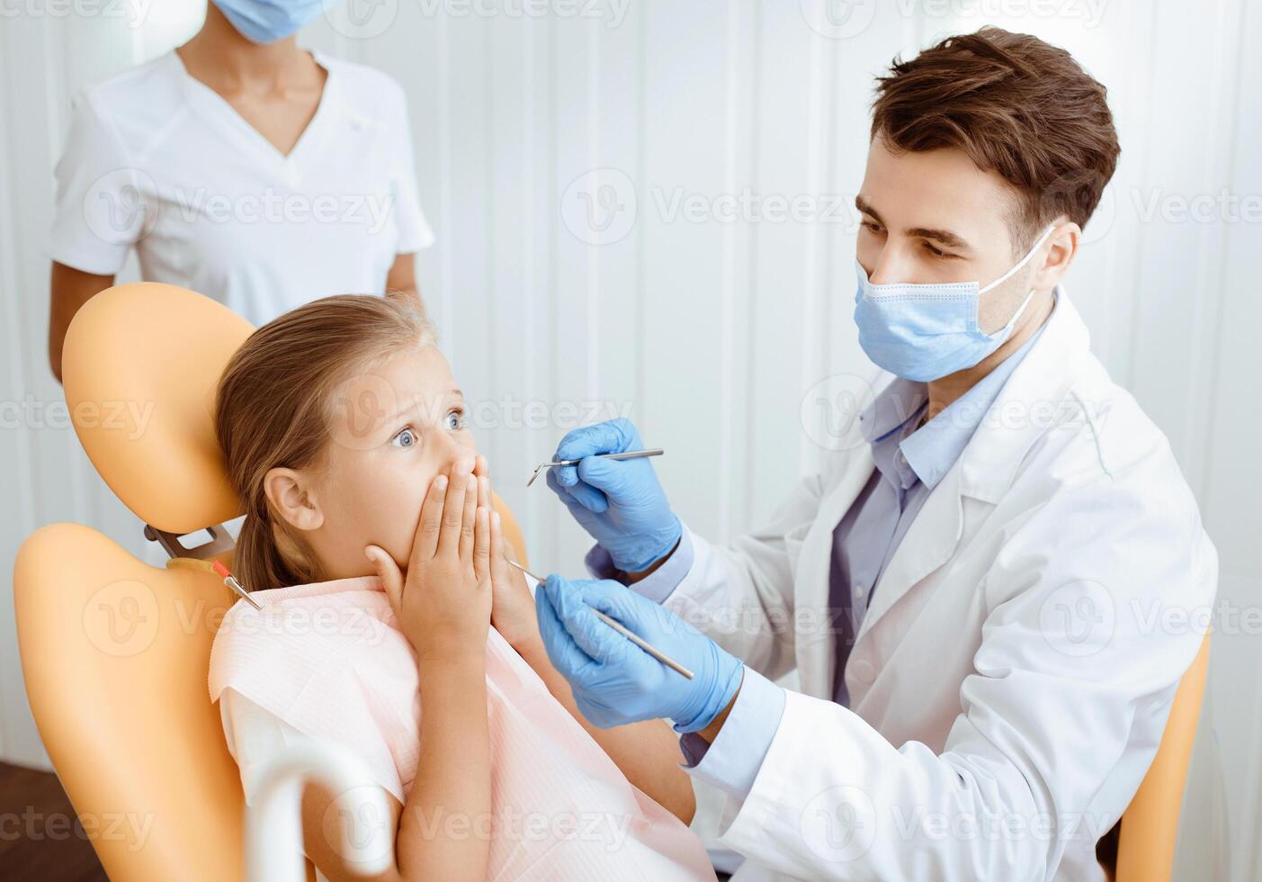 Childrens dentist and work with babies in modern clinic. Scared little girl patient closes mouth and does not allow to doctor in white coat, protective mask and rubber gloves teeth to be treated photo