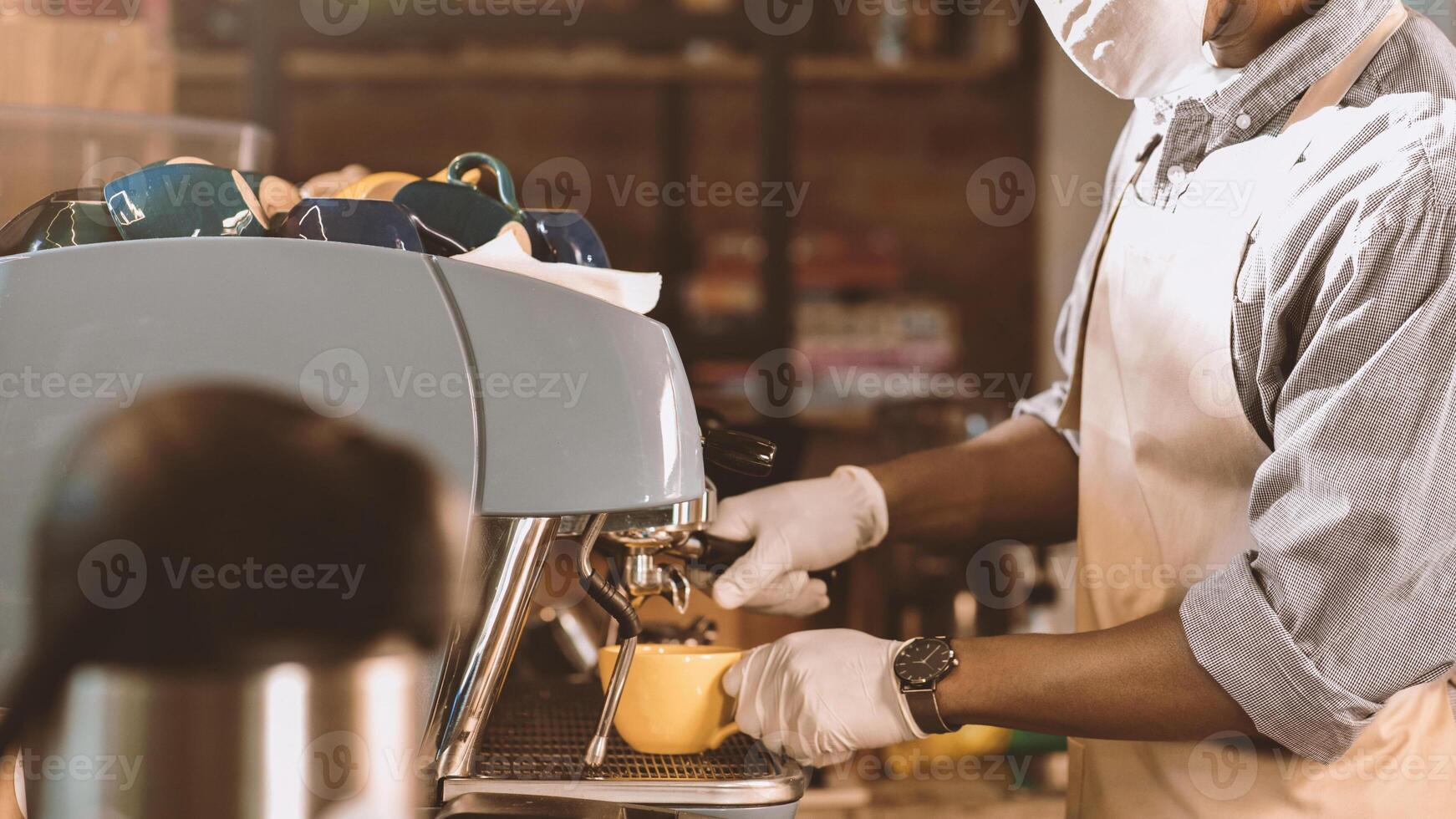 New normal and customer service during covid-19 lockdown. Millennial african american man in apron, protective mask and gloves makes latte in yellow cup on coffee machine at cafe interior, cropped photo