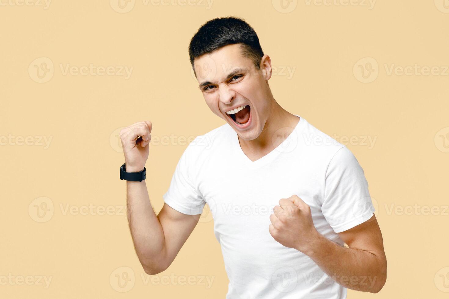 Victory, dance of success, expression of win and great offer. Joyful young man student in white t-shirt shouts and expresses emotions of happy, isolated on beige background, studio shot, free space photo